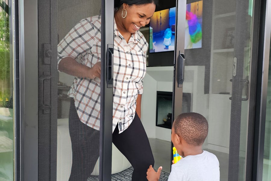A woman is standing in the doorway of her patio door opening a retractable screen. Her two child walking up with excitement.