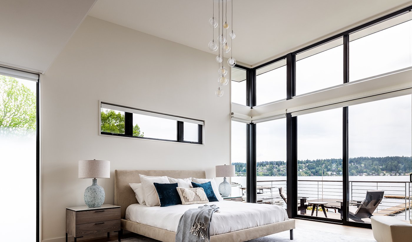 A bedroom with a wall of windows wrapping around a corner and a gliding patio door leading to a balcony with waterfront views.