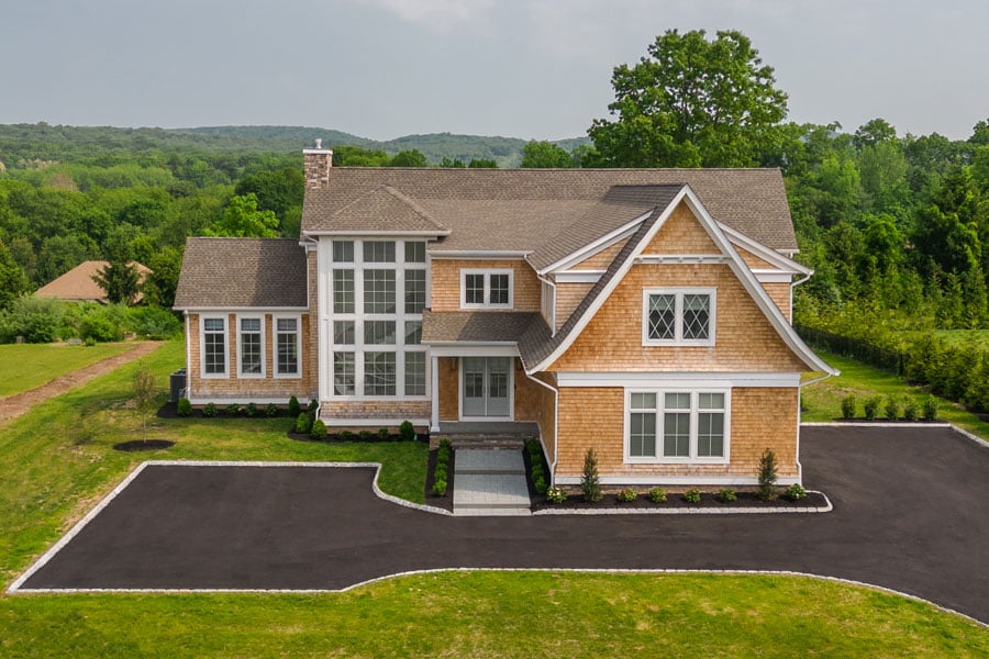 A home with brown wood shingles sits on a large plot of grassy land with a semi-circle driveway. The home has French doors and two stories of white windows with diamond and square window patterns