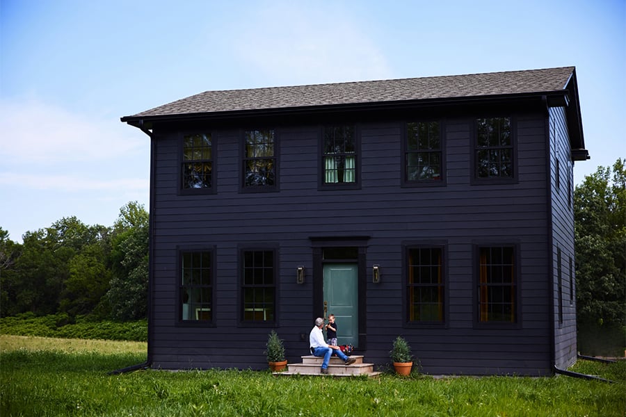 A man and young boy sit on the front steps of a black home with a green front door set in the middle of a grassy field with woods behind.