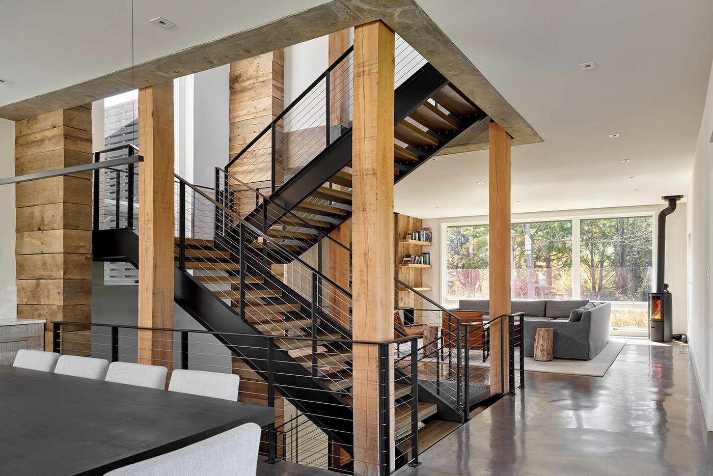An open concept interior space with concrete floors, floor-to-ceiling windows, and an industrial staircase made from a mix of materials, including metal and reclaimed wood.