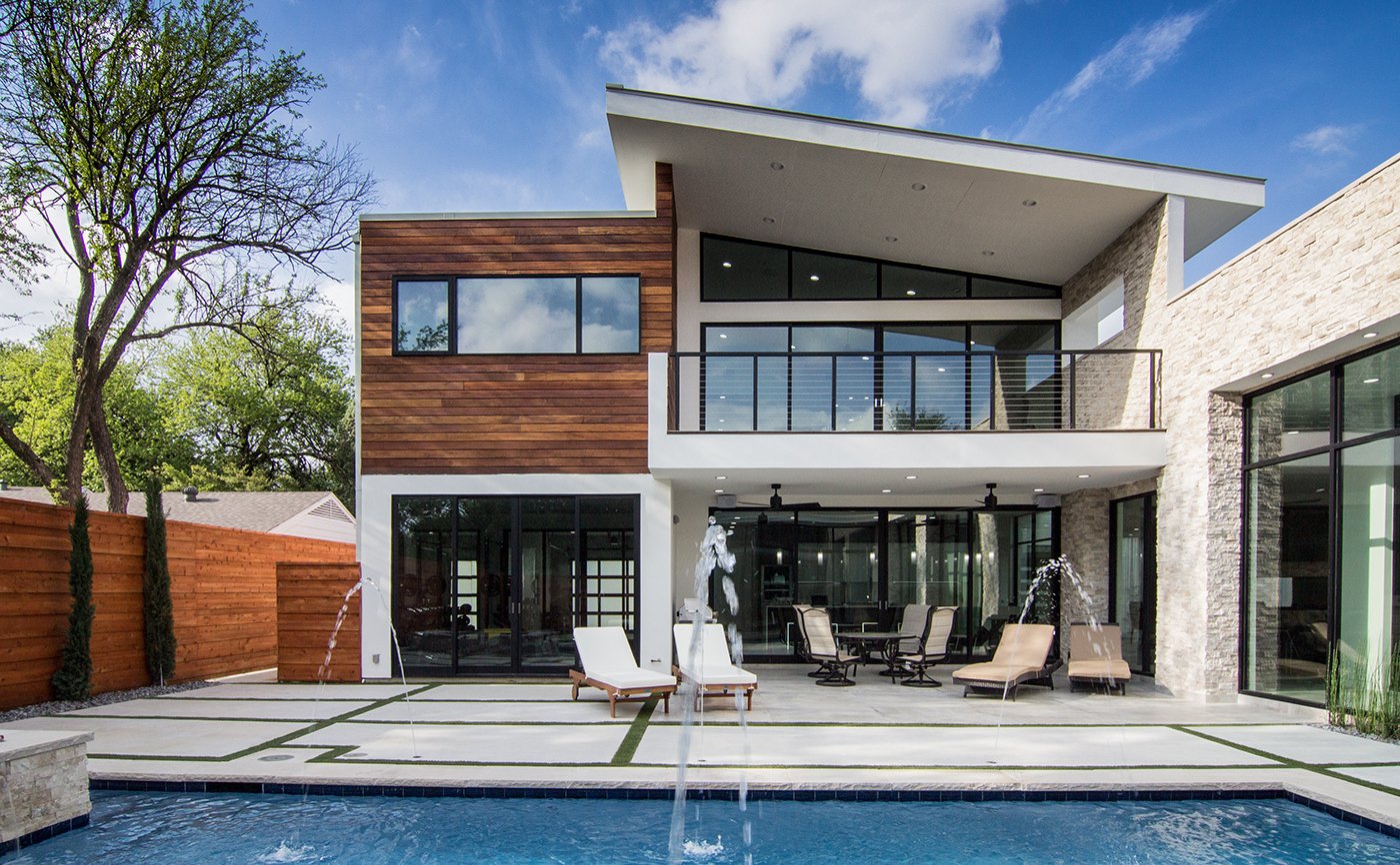 An exterior view of a white and wood modern home with lots of windows and doors facing the swimming pool.