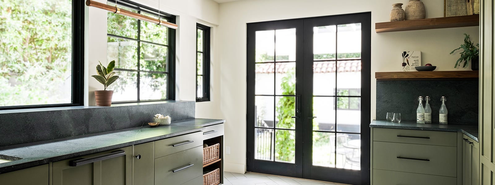 A kitchen with green cabinetry, black soapstone countertops and lots of windows and doors, including a black-framed set of Andersen E-Series Hinged Patio Doors.