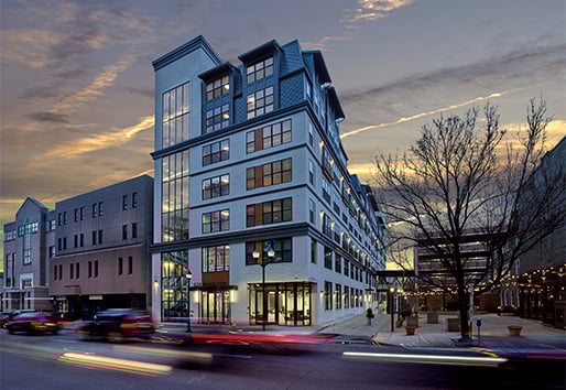 A busy city street with cars driving past an apartment building and plaza. 