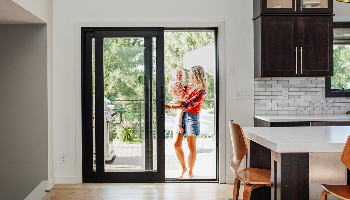 A woman holding a baby opens an Andersen A-Series Gliding Patio Door entering a kitchen from a deck.