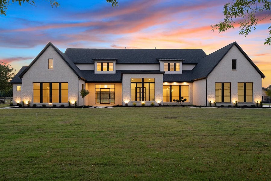 large, ranch style modern farmhouse nestled on a large, well-manicured green lawn. The exterior is a warm white brick color with large black windows with straight line grilles and a black roof that houses three large gables
