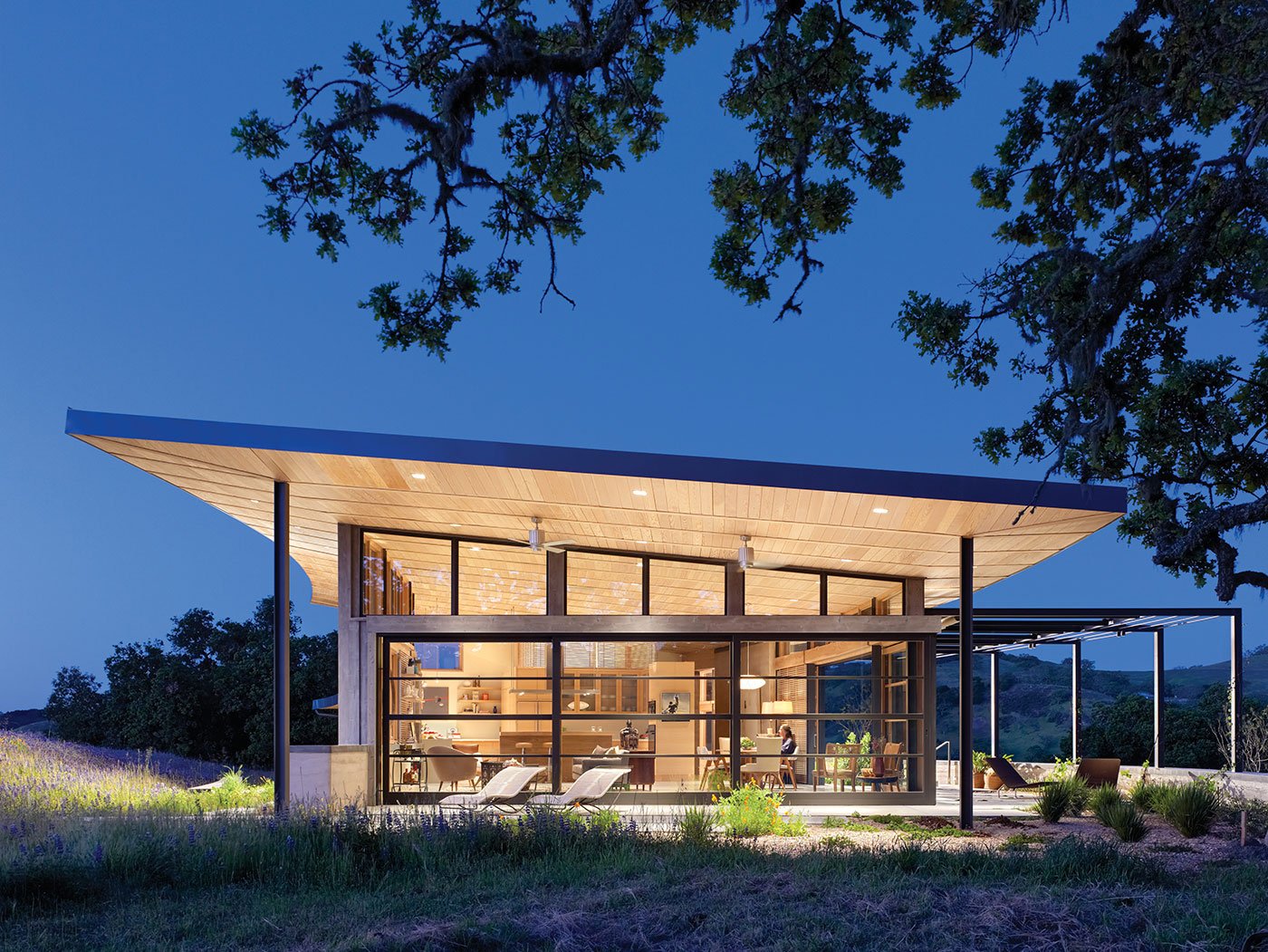 An exterior shot of the home lit up at twilight and viewed from one end so the angle of the roof and glass walls are on display.