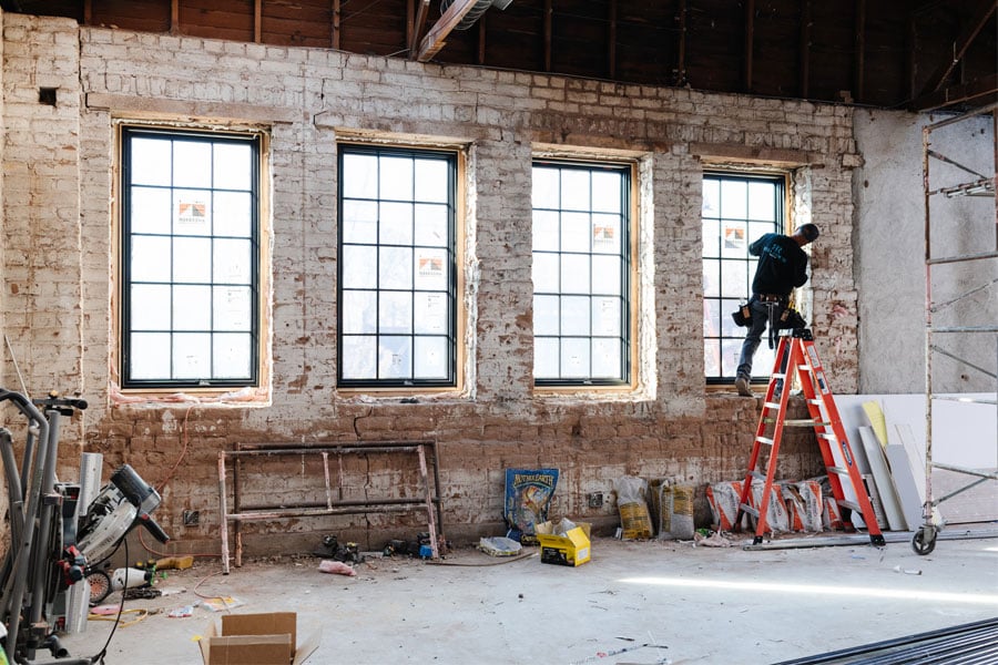 A window installer on a ladder putting the finishing touches on the fourth window in a brick-walled interior.