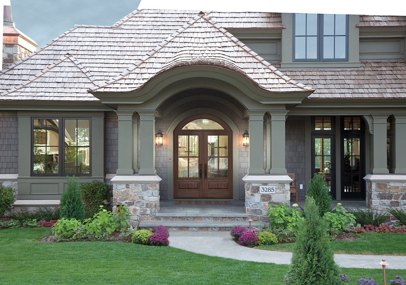 An entryway with a dark wood double front door featuring rectangle glass panels with an arched transom window above, lantern-style lights on either side, and wood shingles roof.