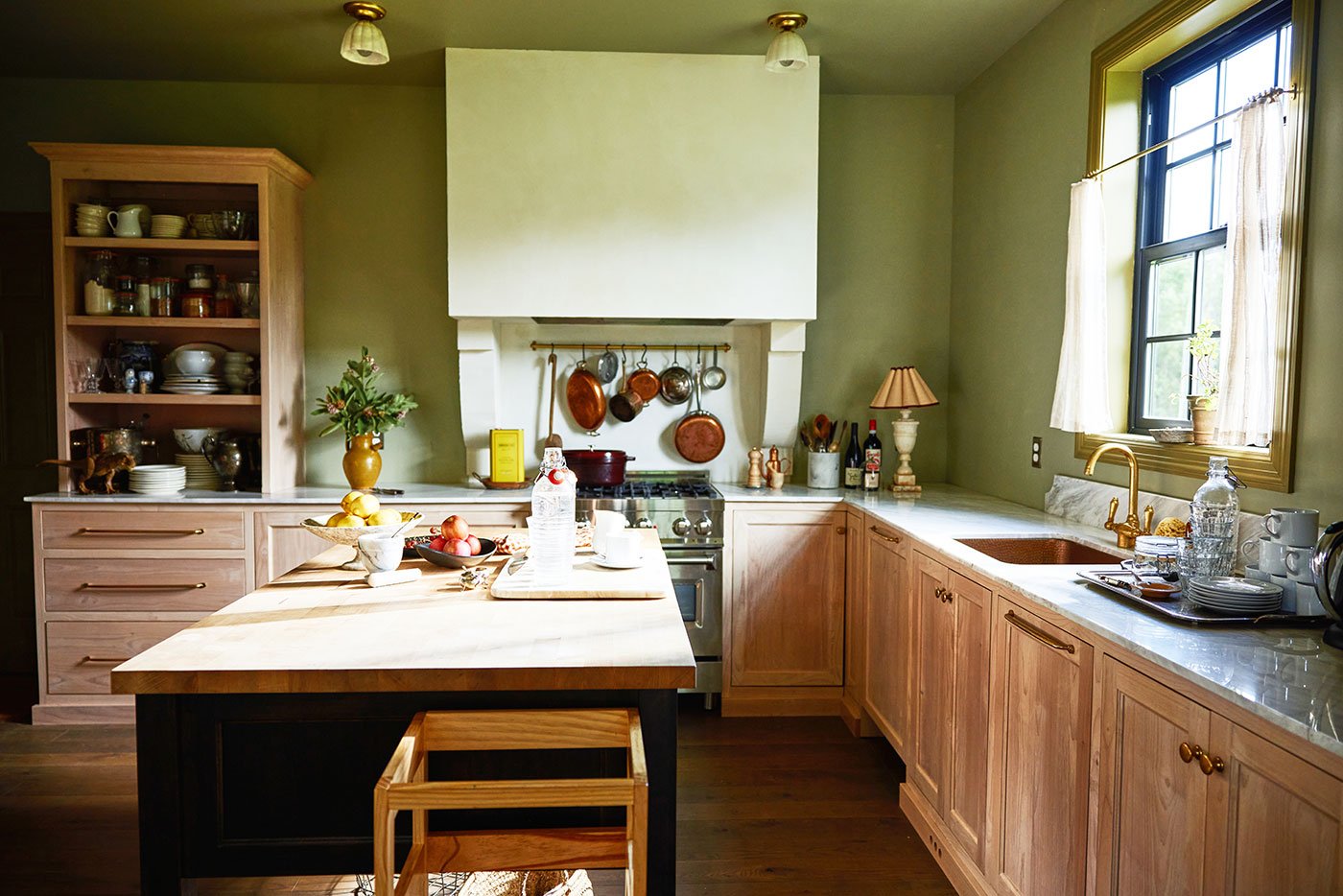 A black single-hung window with a 6-over-6 grille pattern lets a stream of natural light into a cottagecore kitchen featuring an oversized range hood, natural wood cabinetry, and green walls.