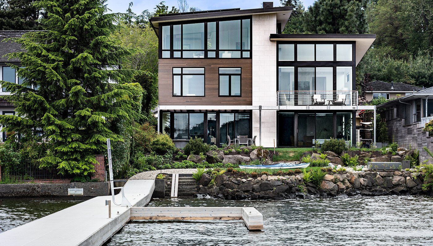 A home next to the water with a white dock, with flat roofs and large windows and patio doors with black frames.