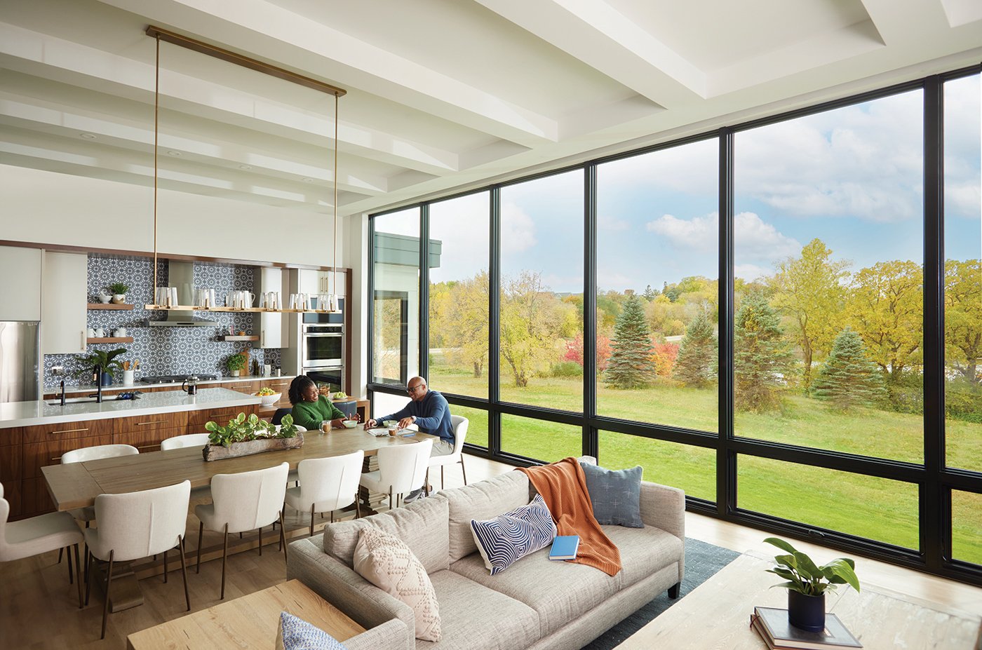 A couple sits at their dining table in an open concept living space that has an entire wall of windows overlooking a green backyard.